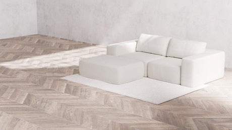 A minimalist living room features a spacious Modular 2-Seater Chaise Sectional in Boucle Aspen, accompanied by a coordinating ottoman on a light beige rug. The herringbone wood floor complements the plain and textured walls, establishing a clean and serene ambiance.