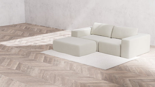 A minimalist room featuring a Modular 2-Seater Chaise Sectional in Boucle fabric, Aspen color, accompanied by a matching ottoman, placed on a textured herringbone wood floor. The furniture rests on a light area rug against a plain white wall, creating a simple and elegant ambiance.