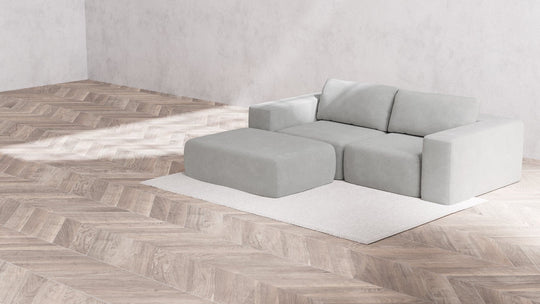 The minimalist living room showcases a Modular 2-Seater Chaise Sectional in Boucle Aspen, perfectly complementing the white rug. The space is enhanced by a herringbone wood floor and plain light gray walls, creating a modern and spacious atmosphere.