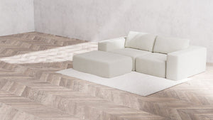 A minimalist living room features the Modular 2-Seater Chaise Sectional in Boucle Aspen complemented by a matching ottoman on a white rug. The floor showcases a herringbone wood pattern, while the plain gray walls contribute to a clean and modern aesthetic.