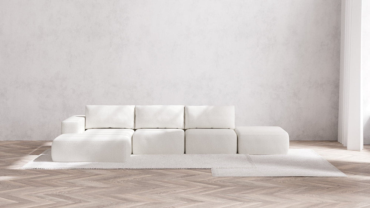 A minimalist living room showcases a white Modular 3-Seater Open-End Chaise Sectional in Boucle Aspen, accentuating clean lines on a light-colored herringbone wood floor. The wall has a textured light gray finish, while a neutral rug enhances the modern, airy aesthetic.