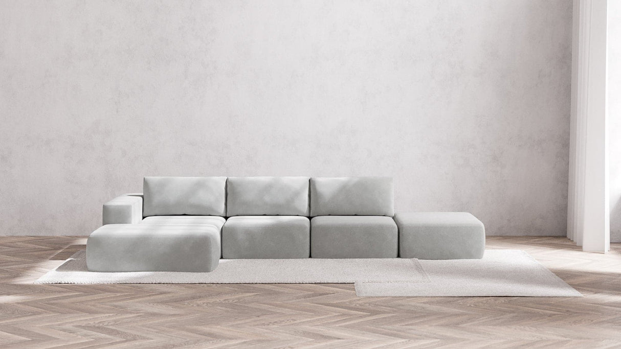 The Modular 3-Seater Open-End Chaise Sectional in Boucle Aspen is placed on a light rug in a minimalist room, featuring light wooden herringbone floors and a textured white wall. Natural light streams in from an unseen window to the right.