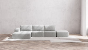 The Modular 3-Seater Open-End Chaise Sectional in Boucle Aspen is placed on a light rug in a minimalist room, featuring light wooden herringbone floors and a textured white wall. Natural light streams in from an unseen window to the right.