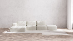 A minimalist living room showcases the Modular 3-Seater Open-End Chaise Sectional in Boucle Aspen, elegantly placed on a light rug. A textured, light gray wall serves as the backdrop, complemented by a herringbone-patterned wood floor to create a clean and modern aesthetic.