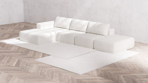 A minimalist living room with light wood herringbone flooring showcases a modern Modular 3-Seater Open-End Chaise Sectional in Boucle Aspen, placed on a white area rug. The walls are unadorned, and the space is bathed in natural light.