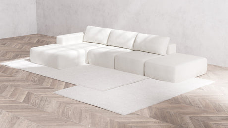 A minimalist living room with light wood herringbone flooring showcases a modern Modular 3-Seater Open-End Chaise Sectional in Boucle Aspen, placed on a white area rug. The walls are unadorned, and the space is bathed in natural light.