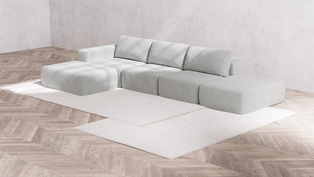 An elegant Modular 3-Seater Open-End Chaise Sectional in Boucle Aspen is arranged on a neutral-toned area rug. The sectional features several square and rectangular pieces, set in a minimalist room with wood-patterned flooring and plain walls.