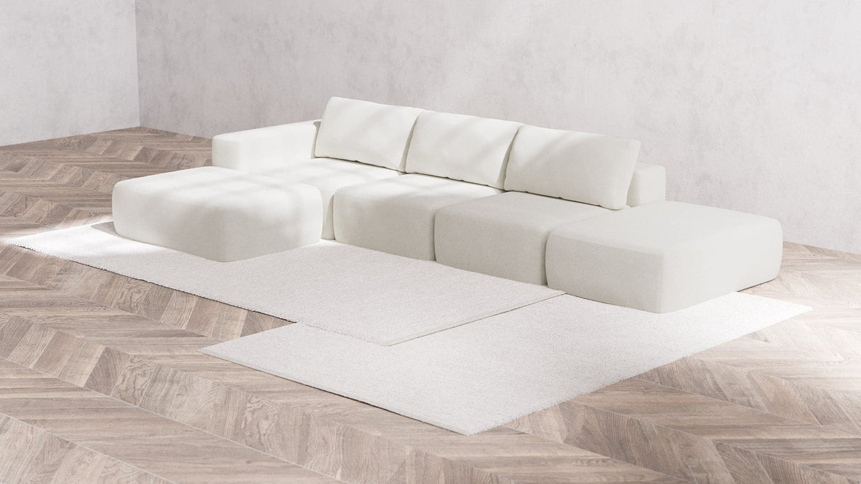 A minimalist living room featuring a Modular 3-Seater Open-End Chaise Sectional in Boucle Aspen, composed of several cushion blocks and backrests. The space is complemented by a light wood herringbone floor and white walls, with a large, light-colored area rug beneath the sofa.