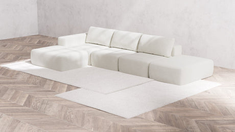 A minimalist living room featuring a Modular 3-Seater Open-End Chaise Sectional in Boucle Aspen, composed of several cushion blocks and backrests. The space is complemented by a light wood herringbone floor and white walls, with a large, light-colored area rug beneath the sofa.