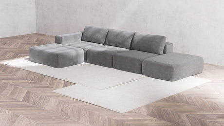 A modern minimalist living room showcases the Modular 3-Seater Open-End Chaise Sectional | Boucle in Aspen, paired with a matching ottoman on a light beige area rug. The room is accentuated by parquet flooring and textured light gray walls, which enhance the sleek and contemporary ambiance.