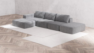 A modern minimalist living room showcases the Modular 3-Seater Open-End Chaise Sectional | Boucle in Aspen, paired with a matching ottoman on a light beige area rug. The room is accentuated by parquet flooring and textured light gray walls, which enhance the sleek and contemporary ambiance.