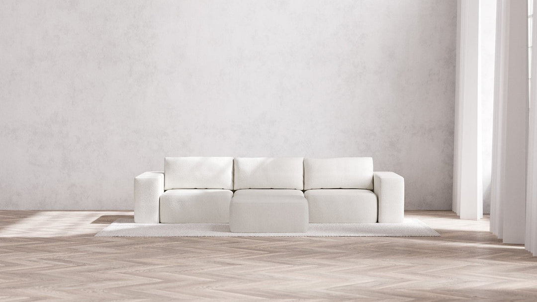 A minimalist, modern living room showcases the Modular 3-Seater Sofa + Ottoman in Boucle Aspen placed on a light area rug. The space is complemented by neutral-toned walls and herringbone-patterned wooden flooring, with ample natural light streaming through large windows.