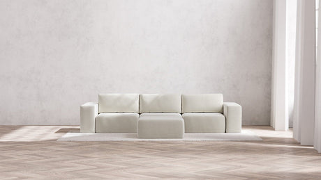 In a bright, spacious room with a light wood herringbone floor and large windows flooding the space with natural light, the Modular 3-Seater Sofa + Ottoman in Boucle Aspen sits gracefully on a textured rug. It features four square sections and is paired with a matching ottoman, creating an elegant minimalist look.