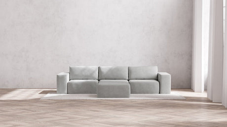 A minimalist, modern living room featuring the light gray Modular 3-Seater Sofa + Ottoman in Boucle Aspen atop a textured rug, set against a plain gray wall. Natural light pours in through tall windows, highlighting the herringbone-patterned wooden floor.
