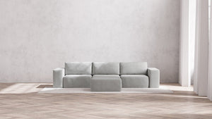 A minimalist, modern living room featuring the light gray Modular 3-Seater Sofa + Ottoman in Boucle Aspen atop a textured rug, set against a plain gray wall. Natural light pours in through tall windows, highlighting the herringbone-patterned wooden floor.