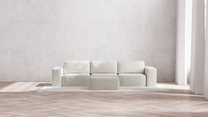 A minimalist living room showcases a Modular 3-Seater Sofa and Ottoman in Boucle Aspen, set against a light gray wall. The room is illuminated by natural light streaming through a large window, highlighting the textured wooden floor. Beneath the sofa sits a white rug.