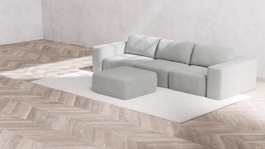 A minimalist living room is styled with the Modular 3-Seater Sofa and Ottoman in Aspen Boucle on a white rug, complemented by light wood herringbone flooring and neutral-toned walls for a clean, modern aesthetic.