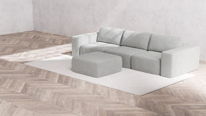 A minimalist living room is styled with the Modular 3-Seater Sofa and Ottoman in Aspen Boucle on a white rug, complemented by light wood herringbone flooring and neutral-toned walls for a clean, modern aesthetic.