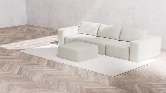A minimalist room featuring a large Modular 3-Seater Sofa and matching Ottoman in Boucle Aspen on a textured white rug. The floor showcases a light wood herringbone pattern, and the plain white walls contribute to a clean, modern aesthetic.