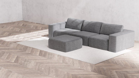 In the minimalist living room, a large Modular 3-Seater Sofa and Ottoman in Boucle Aspen sits atop a light beige rug. The herringbone-patterned wooden flooring complements the plain gray walls. The space is well-lit, resulting in a cozy and modern atmosphere.