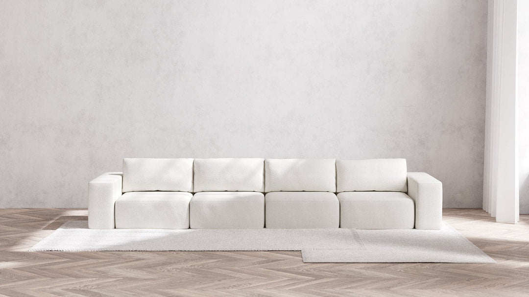 A minimalist living room showcases a Modular 4-Seater Sofa | Boucle in Aspen, characterized by its long and modular design with square cushions, elegantly placed against a textured light gray wall. The wooden herringbone floor is partially adorned with a light gray area rug.