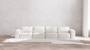 A minimalist living room showcases a Modular 4-Seater Sofa | Boucle in Aspen, characterized by its long and modular design with square cushions, elegantly placed against a textured light gray wall. The wooden herringbone floor is partially adorned with a light gray area rug.