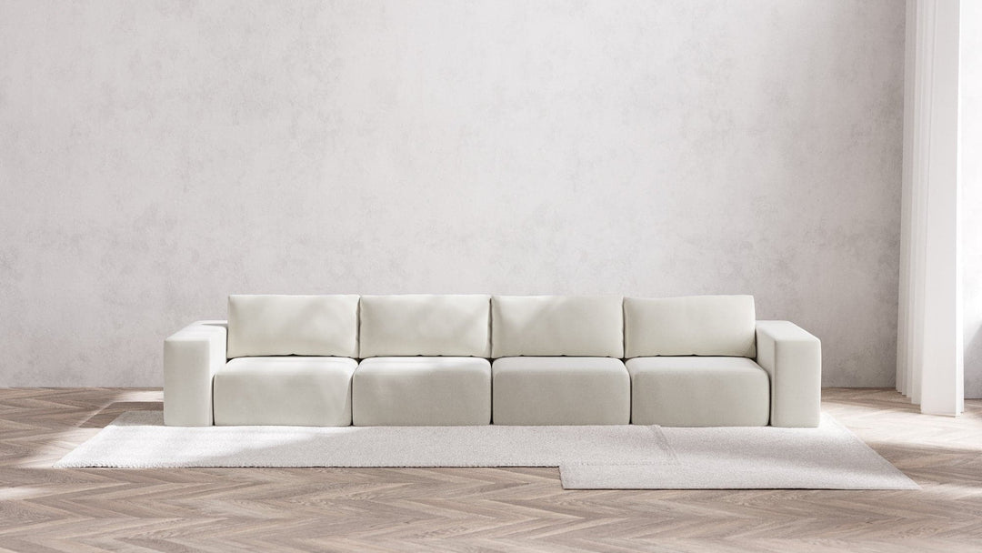 A minimalist room showcasing a large, modern Modular 4-Seater Sofa in Boucle Aspen that features square cushions and block armrests. The floor is adorned with a chevron pattern, and a light carpet peeks out from beneath the sofa. The walls are plain and light-colored.