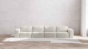 A minimalist room showcasing a large, modern Modular 4-Seater Sofa in Boucle Aspen that features square cushions and block armrests. The floor is adorned with a chevron pattern, and a light carpet peeks out from beneath the sofa. The walls are plain and light-colored.