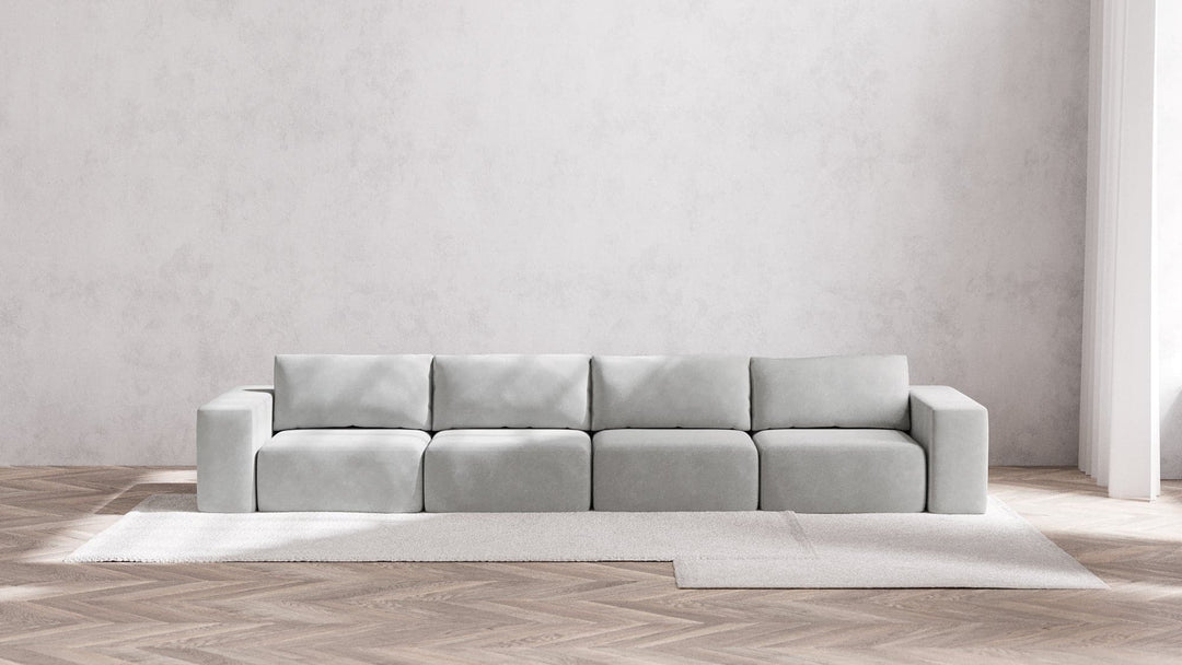 A minimalist living room featuring the Modular 4-Seater Sofa in Boucle Aspen set against a plain white wall. The light gray sofa is placed on a beige rug laid out on herringbone-patterned wooden flooring.