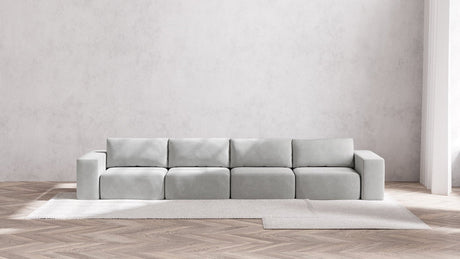 A minimalist living room featuring the Modular 4-Seater Sofa in Boucle Aspen set against a plain white wall. The light gray sofa is placed on a beige rug laid out on herringbone-patterned wooden flooring.