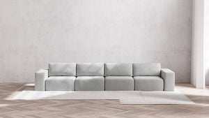 A minimalist living room featuring the Modular 4-Seater Sofa in Boucle Aspen set against a plain white wall. The light gray sofa is placed on a beige rug laid out on herringbone-patterned wooden flooring.