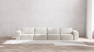 A minimalist room with a Modular 4-Seater Sofa in Boucle Aspen positioned against a light gray wall. The floor displays a herringbone wood pattern, topped with a large, light-colored rug. Natural light streams in from an unseen window.