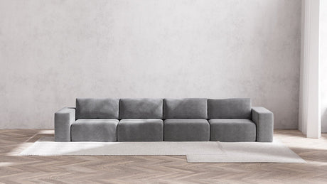 A modern, minimalist living room showcases the Modular 4-Seater Sofa in Boucle Aspen, positioned against a light gray wall. The sofa rests atop a large, light-colored area rug laid over a wooden herringbone floor, cultivating a serene and airy ambiance.