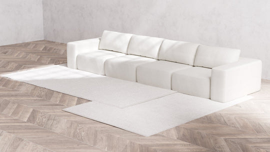 A minimalist room showcases the long Modular 4-Seater Sofa in Boucle Aspen on a light wood herringbone floor. Two matching rectangular white rugs are partially layered in front of the sofa, while the plain, light-colored walls enhance the room's simplicity.