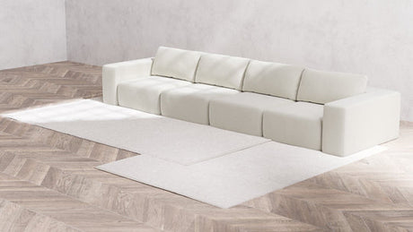 A modern, minimalist living room showcases the Modular 4-Seater Sofa in Boucle Aspen along with five cushions against a light gray wall. The floor features a light wood chevron pattern, and two white rugs are elegantly positioned in front of the sofa.