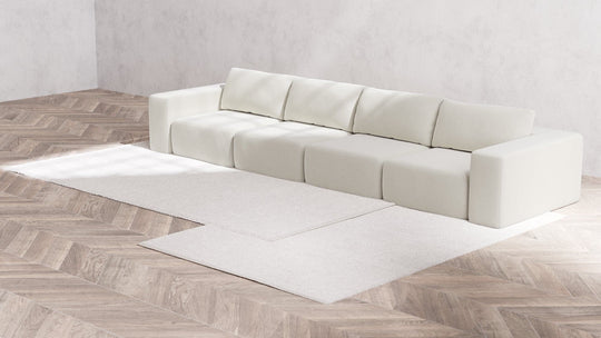 A modern, minimalist living room showcases the Modular 4-Seater Sofa in Boucle Aspen along with five cushions against a light gray wall. The floor features a light wood chevron pattern, and two white rugs are elegantly positioned in front of the sofa.