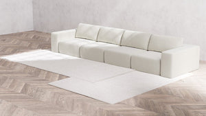 A modern, minimalist living room showcases the Modular 4-Seater Sofa in Boucle Aspen along with five cushions against a light gray wall. The floor features a light wood chevron pattern, and two white rugs are elegantly positioned in front of the sofa.