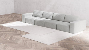 A Modular 4-Seater Sofa in Boucle Aspen stands against a textured light gray wall. It rests on a light-colored herringbone-patterned floor, complemented by two overlapping rectangular rugs in a harmonious neutral tone.