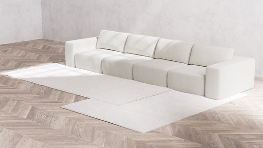 A minimalist room features the Modular 4-Seater Sofa in Boucle Aspen, positioned against a light gray wall. The sofa rests on a light wooden herringbone floor, which is partially covered by two overlapping cream-colored rugs.