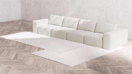 A minimalist room features the Modular 4-Seater Sofa in Boucle Aspen, positioned against a light gray wall. The sofa rests on a light wooden herringbone floor, which is partially covered by two overlapping cream-colored rugs.