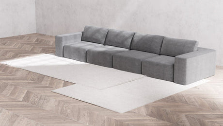 A modern, Modular 4-Seater Sofa in Boucle Aspen sits in a minimalist living room with wooden herringbone flooring. Two white, rectangular rugs are placed partially under the sofa. The walls are bare and white.