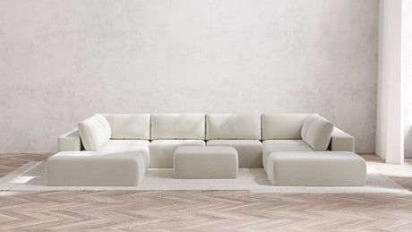 In a modern, minimalist living room, a large Modular 8-Seater Open-Ends U Sectional and matching Ottoman in Boucle Aspen take center stage on a clean white rug. The space maintains its neutral color palette, highlighted by a herringbone wooden floor and textured white walls. Soft natural light streams in from the right.