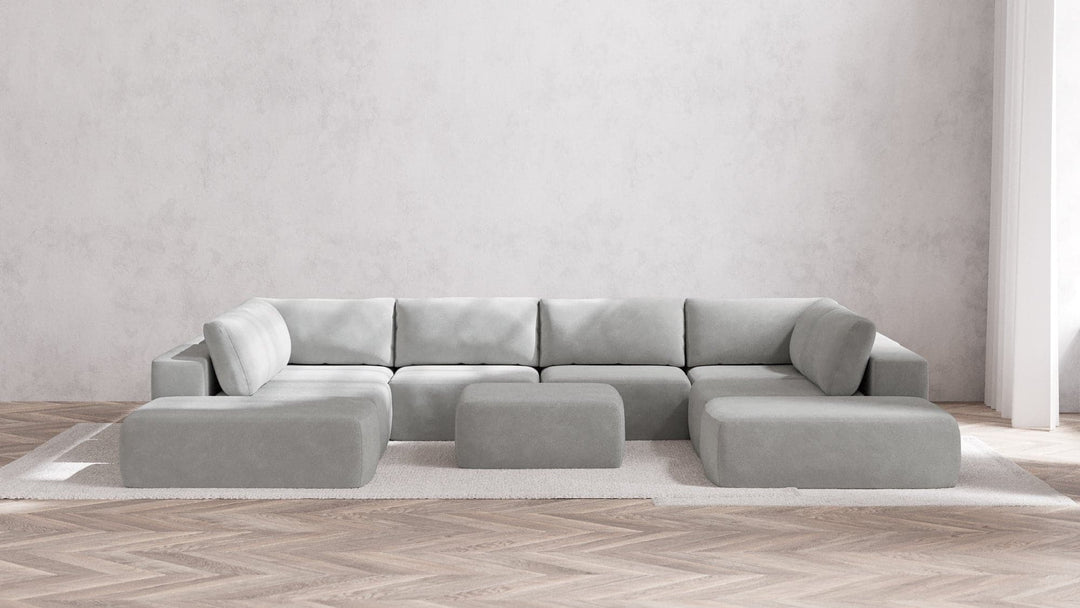 A spacious living room showcases a modern Modular 8-Seater Open-Ends U Sectional + Ottoman in Boucle Aspen, arranged on a white rug. The room embraces a minimalist design with pale, textured walls and wooden herringbone flooring.
