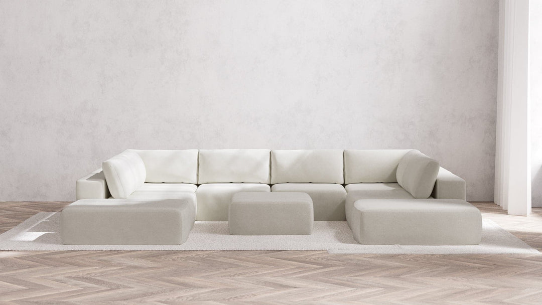 A minimalist living room showcases the Modular 8-Seater Open-Ends U Sectional + Ottoman in Boucle Aspen, featuring clean lines and a modern design. This ensemble sits on a white rug atop a light herringbone wooden floor, set against a plain white wall.