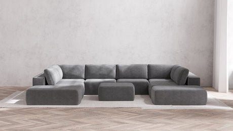 A minimalist living room is highlighted by the Modular 8-Seater Open-Ends U Sectional + Ottoman in Boucle Aspen, which boasts sleek modern lines. Two matching gray ottomans complement the arrangement, all set on a light-colored area rug atop herringbone wood flooring, framed by a plain white wall.