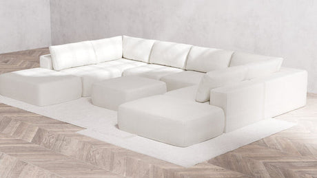 A minimalist living room showcases the Modular 8-Seater Open-Ends U Sectional + Ottoman in Boucle Aspen, providing multiple sections and cushions. The arrangement rests on a light wooden herringbone floor atop a white rug, set against a plain gray wall.