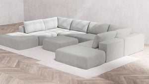A contemporary, minimalist corner sofa styled as the Modular 8-Seater Open-Ends U Sectional + Ottoman in Boucle Aspen graces a white rectangular rug. It features a modular design accompanied by matching ottomans. The flooring showcases light herringbone wood, complemented by plain gray walls.