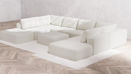 A large, contemporary white "Modular 8-Seater Open-Ends U Sectional + Ottoman | Boucle in Aspen" with firm cushions is placed in the corner of a room featuring light wood herringbone flooring. The sectional rests on a white rug, fostering a minimalist and cozy ambiance.