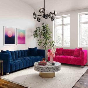 A modern living room features a Bea Velvet Tufted Sofa in blue and another in pink, positioned to face each other. A round coffee table adorned with books and pink flowers rests on a beige rug. On the wall hang two abstract artworks, while a large plant stands by the window.