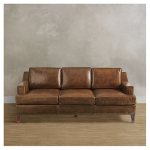 The Collins Sofa, a polished brown leather three-seater, is placed against a neutral, textured gray wall on a wooden floor. It features cushion backrests and armrests, adding a classic and elegant look to the room.